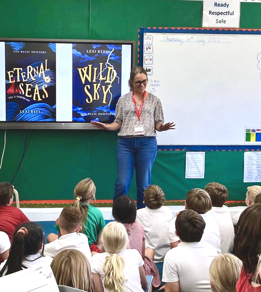 Lexi Rees doing a school assembly with students seated and two of her book covers on the screen behind